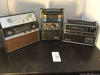 Three vintage portable radios placed side by side on a black surface showing front panels, knobs, handles, and frequency displays.