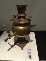 Photo of an antique brass samovar with spout, handles, and base, on a white surface.