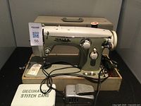 Photo showing the White brand vintage sewing machine placed in front of its beige hard carrying case. Included are sewing machine power cord, foot pedal, and a box labeled Decorative Stitch Cams on the base.