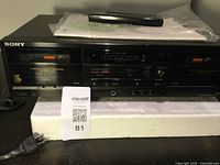 Front view of Sony cassette player recorder showing dual cassette decks labeled Deck A and Deck B, control buttons, analog VU meter, power cord, and remote control on top.