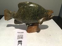 Full profile view of stone salmon sculpture on white support surface, showing carved details and signature.