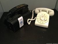 Photo showing a black vintage portable automobile telephone with push-button keys alongside a white rotary dial home phone with coiled cord and handset.