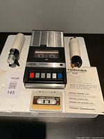 Vintage Toshiba KT-1500 cassette recorder/player with mechanical control buttons and metal speaker grille top. Two external microphones, one white and one black, and cassette tape with label. User manual included.