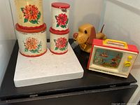 Photo showing stacked tin boxes with floral patterns and the yellow wind-up toy dog behind them, also visible is the Fisher-Price toy on the right.