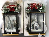 Front view of two Thomas Kinkade holiday lantern decorations with Christmas greenery and red bows on top, showing interior village scenes with snow and figurines