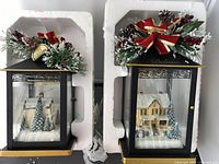 Two black metal Thomas Kinkade winter-themed lanterns in their styrofoam packaging, showing festive greenery topped with red ribbon bows and detailed snowy village scenes behind glass panels.