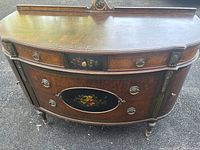 Front view of antique sideboard showing curved shape, wood finish, metal ring handles, and hand-painted floral design on drawers.