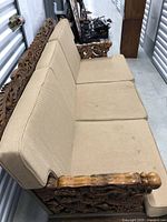 Side-angle photo of the wooden sofa with beige cushions, showing full length and carved wooden arms.