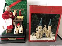 Photo of holiday figurine scene showing character in red, table with parchment and money bags, and original packaging in background.
