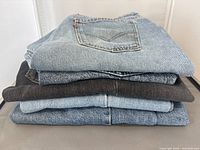 Stack of five folded denim items showing both light blue and black colors, stacked with the light blue Levi's brand back pocket visible on top