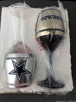 Two Dallas Cowboys themed wine glasses in foam packaging, one with star logo, the other with COWBOYS text, both wrapped in plastic.