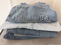 Stack of folded medium blue denim clothing including pants and shorts, showing back pockets and denim texture.
