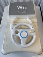 Wii accessories and manual on white surface: white Wii Steering Wheel, white Controller Nunchuk, and Wii Operations Manual with grey cover.