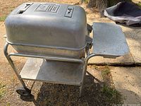 Full view of the aluminum grill showing closed hinged lid, attached side shelf, and lower storage shelf on wheels and legs.