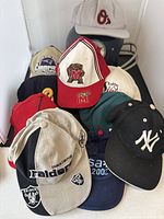 Front view of 10 hats with various team logos arranged in stack, with black football helmet behind.