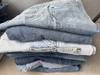 Stack of ten pairs of assorted medium blue denim jeans showing variety of blue shades and denim textures, view from folded side and back pockets visible.
