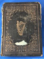 Front cover shows black leather with an ornate gold embossed border and central harp emblem with the name 'John Ellis'. Visible wear, scuffing, and some damage to lower part of cover.