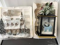 Photo showing two holiday decorations in original styrofoam packaging: a snow-covered church and a black lantern with winter house scene