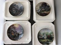 Photo showing all 4 plates placed individually in foam holders, each with distinct scenic artwork typical of Thomas Kinkade's style.