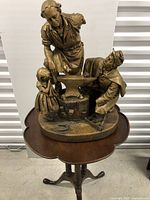 Side table with twist leg design and brown finish, topped by resin sculpture of blacksmith scene with missing arm on one figure