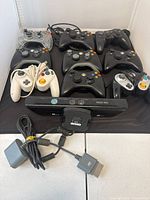 Photo of 10 console controllers and Kinect sensor placed on black surface showing both wired and wireless types, including Xbox 360 and Nintendo GameCube models.