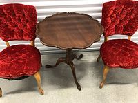 Two red velvet upholstered chairs with tufted backs positioned on either side of a scalloped edge mahogany round table with three curved legs. Minor surface scratches visible on table.
