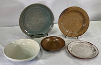 Five ceramic pottery items including three plates, one bowl, and one dish arranged on a surface, varying in color from gray-green, brown, off-white, and speckled white-brown.