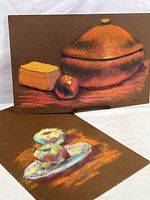 Photo showing two pastel still life paintings on brown paper, one with a covered dish, cheese block, and apple, and the other with pastries on a plate.