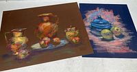 Two original pastel still life paintings laid flat, one showing two pitchers with fruit and a covered dish on a brown background, the other with a blue pot and apples on dark background.