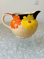 Side view of ceramic pitcher showing orange and yellow floral design with beige body and black and orange trim on handle and spout.