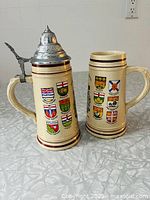 Pair of Canadian Centennial ceramic steins showing front sides with provincial crests and brown ring accents. One stein has a metal lid with thumb lever.