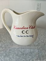 Front side of cream-colored ceramic Canadian Club whisky pitcher showing red 'Canadian Club' text, black 'CC' with gold accent, and blue slogan 'The Best In The House'.