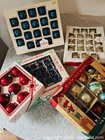 Five boxes of Christmas ornaments on a silver textured table including blue, red, gold, and assorted colors with original boxes.
