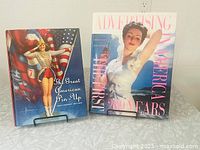 Two coffee table books displayed upright on metal stands showing covers: The Great American Pin-Up with a patriotic pin-up girl and Advertising America with a vintage advertisement featuring a woman.