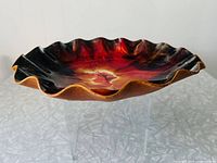 Photo showing side angle of round dish with scalloped edges and vibrant fiery color palette in red, black, and yellow-orange hues, displayed on clear stand.