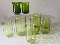 Nine green glasses arranged on a table. Includes three taller olive green Vertical Optic Wave glasses, four shorter matching optic wave glasses, and two smaller curved green juice glasses.
