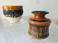 Both pottery pieces shown side by side on a textured white surface. The cup is on the left with a cream and amber brown glaze and bubbled texture. The vase is on the right featuring vertical textured stripes in brown, black, and orange with a flared brown rim.