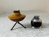 Pair of table lighters shown side by side on a light gray textured surface. The larger tripod lighter is brass-colored with three angled legs, smaller lighter is rounded with a dark transparent body and chrome top.