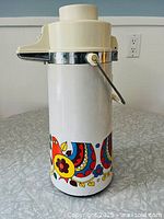 Full view of tall cylindrical white beverage dispenser with colorful Scandinavian bird and floral motifs on lower half, plastic top with pump and chrome band, and handle attached.
