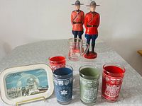 Full lot view showing four Canadian Centennial glasses, two plastic Mountie figurines, the Expo67 metal tray, and commemorative shotglass.