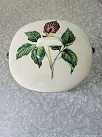 Top view of the white lid of the casserole dish showing detailed green leaves and single purple flower design with handle.