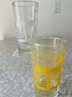 Pair of vintage Dominion Glass Company cocktail glasses with printed graphics and cocktail recipes, one with yellow print and one with white print, shown on white stand.