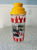Front side of the vintage Medco glass cocktail shaker showing yellow plastic lid and barbershop singer graphics.