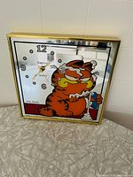 Square mirrored glass wall clock with gold-tone frame featuring Garfield brushing his teeth illustration, black Arabic numerals, yellow clock hands, STAMFORD Quartz branding, illustration by Jim Davis.