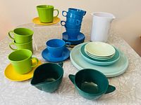 Photo of the full set showing multi-colored plastic kitchenware including cups, plates, bowls, creamer, sugar bowl, and pitcher arranged on a table with a patterned tablecloth.