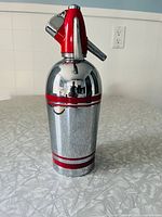 Full view of the vintage soda syphon showing its chrome body, red plastic top with dispensing lever, and red horizontal stripes.