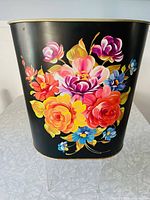 Front view of black oval metal waste bin showing colorful floral illustration with large pink, yellow, orange, red, blue, and purple flowers and green leaves.