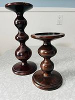 Two polished contemporary wood candle stands shown side by side, dark finish, rounded sculpted design with bulbous forms and circular bases and tops.
