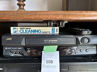 Front view of JVC VHS player with Fuji Video Head Cleaning Cassette on top and remote control on the right side.