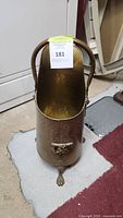 Large brass umbrella stand with curved handle and claw feet shown from front on carpeted floor near door